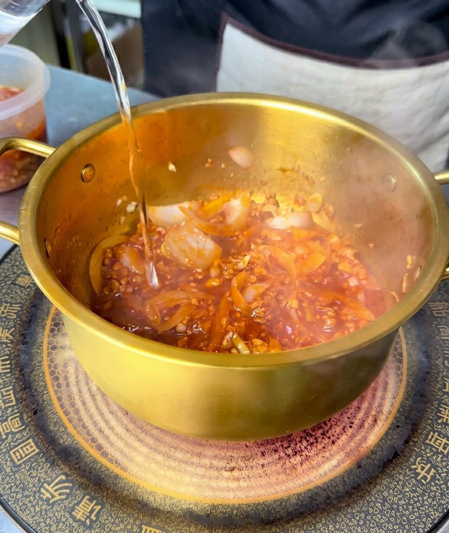 Clear water being poured into a golden pot containing a red, spicy stir-fried meat and vegetable base.