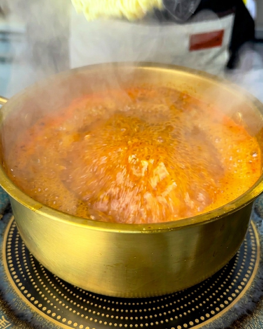 A block of dried instant ramen noodles being submerged into a pot of bubbling red soup.