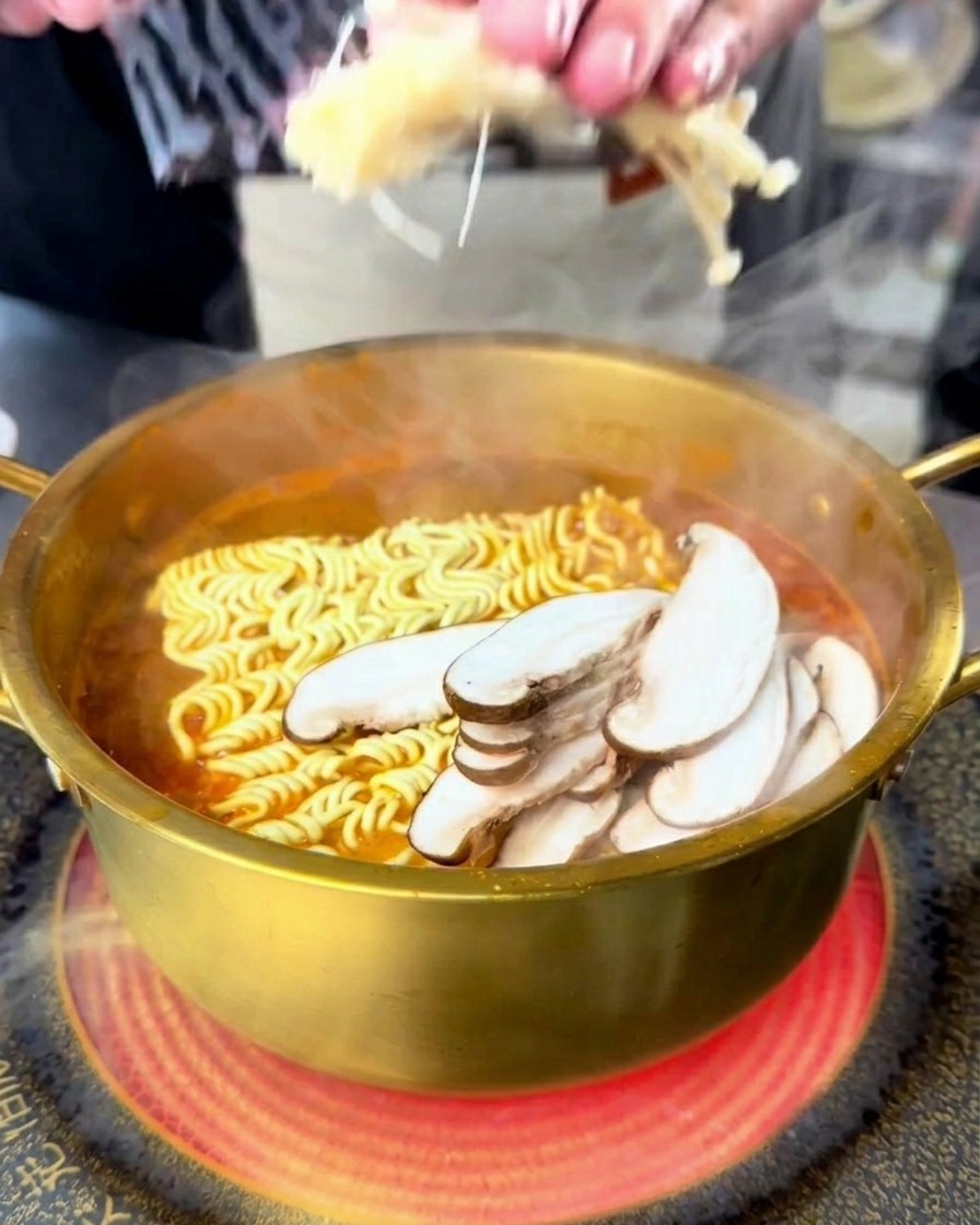 A person adding enoki and sliced shiitake mushrooms to a boiling pot of seafood ramen.