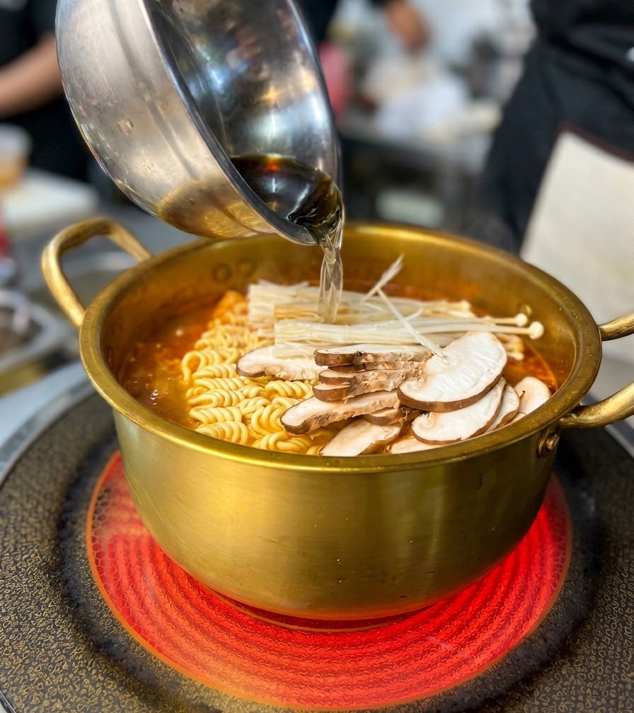 Adding a bowl of sliced mushrooms and seafood into a golden pot filled with ramen noodles and a red spicy broth.