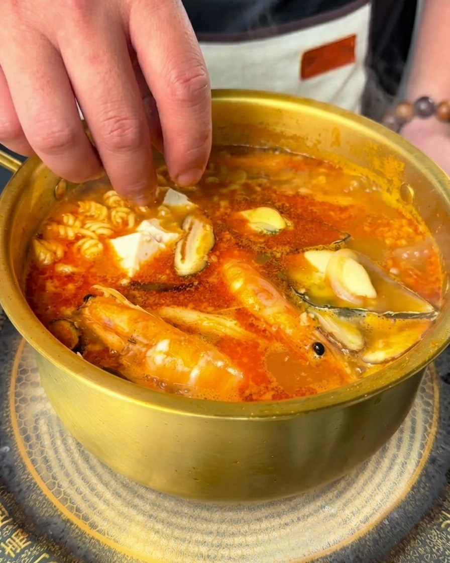 Hand dropping white cubes of soft tofu and fresh chili slices into a pot of bubbling spicy seafood ramen.