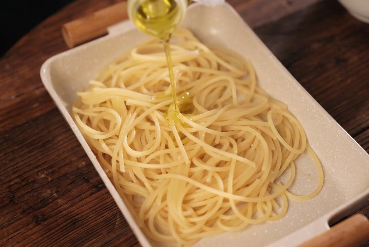 Cooked spaghetti in a cream-colored rectangular tray being drizzled with yellow olive oil from a bottle.