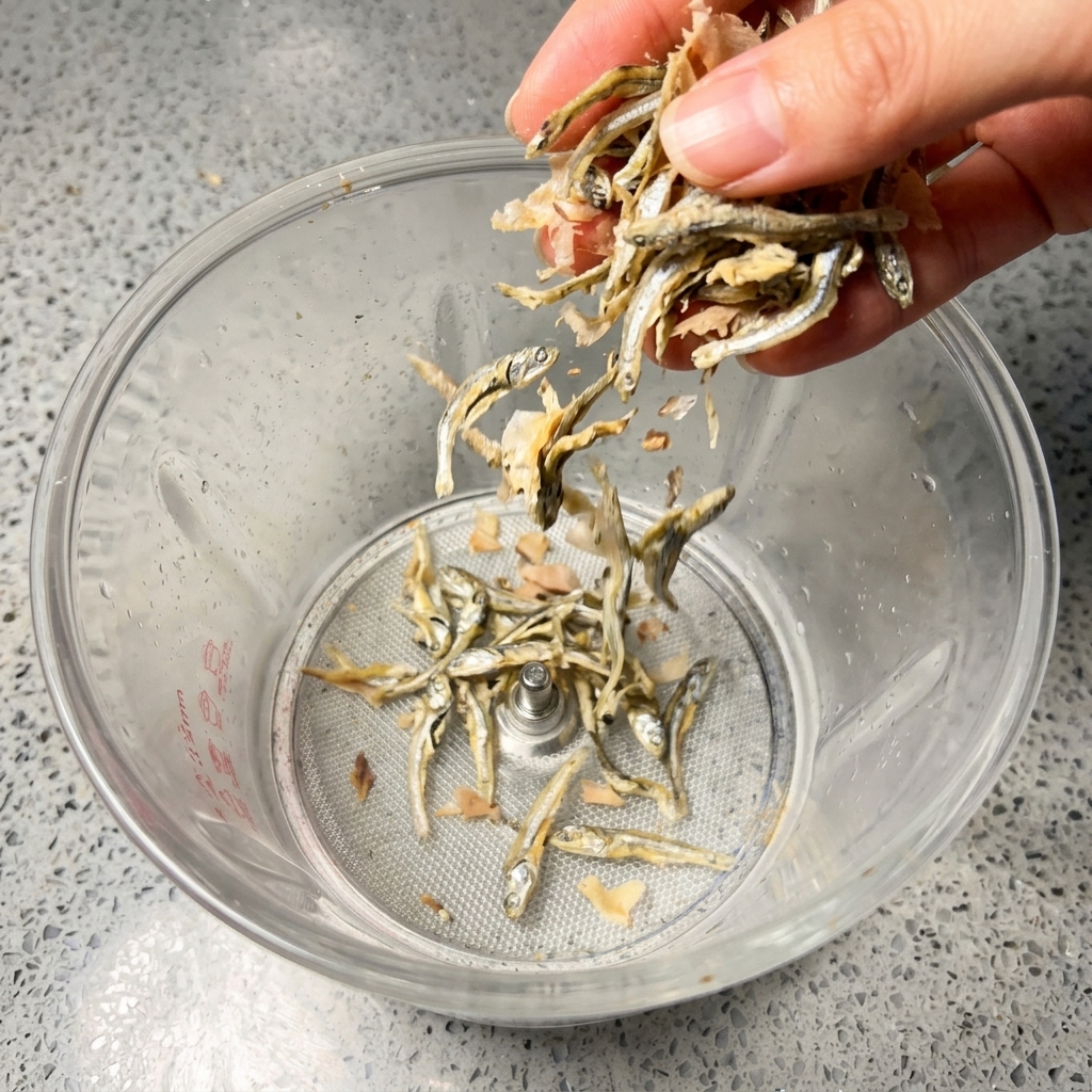 A hand dropping small dried silver anchovies and pinkish-brown bonito flakes into a clear blender jar.