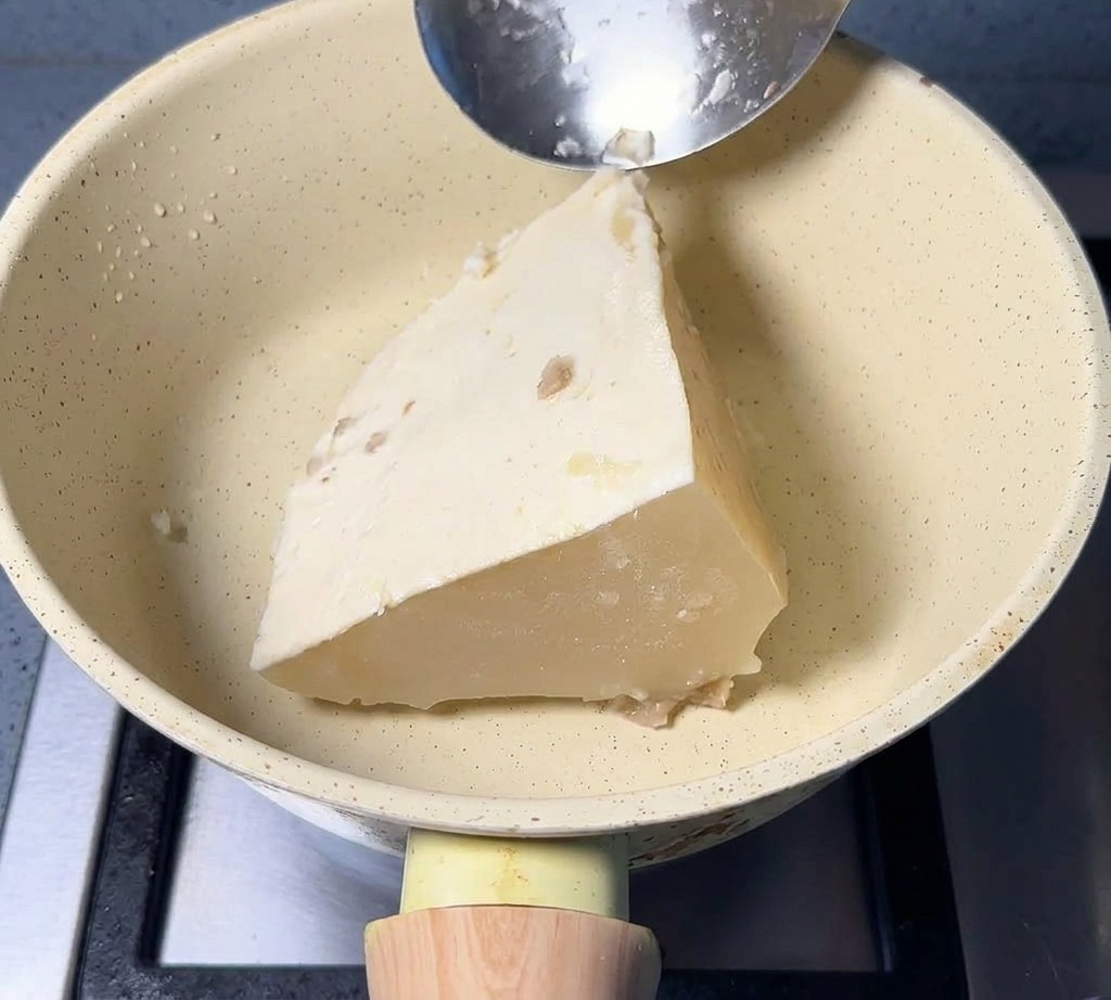 A large triangular block of frozen white pork broth sitting in a cream-colored pan with a metal spoon resting on the edge.