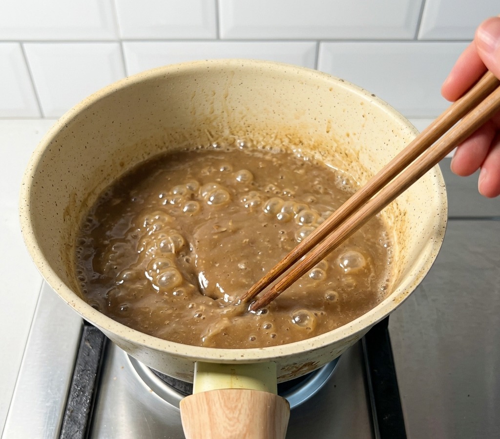 A small beige pot on a gas stove containing boiling brown pork and fish broth being stirred with wooden chopsticks.