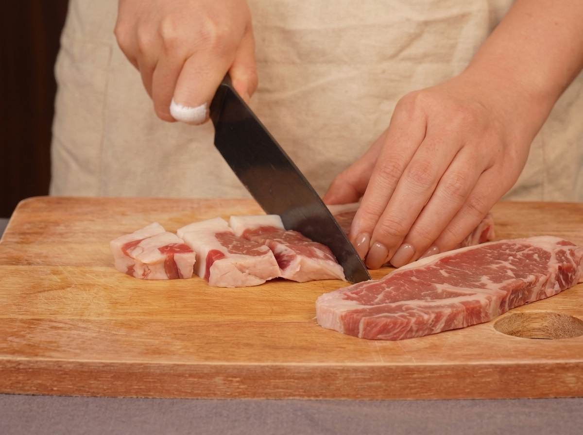 A person using a chefs knife to slice raw marbled beef into chunks on a wooden cutting board.