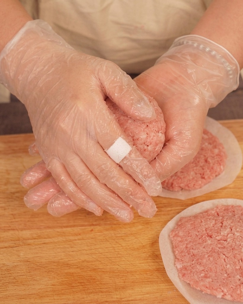 A person wearing clear plastic gloves tossing a ball of raw ground beef between their hands over a wooden cutting board.