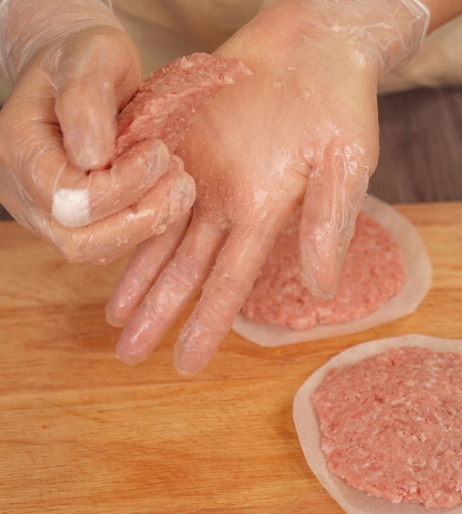 Gloved hands pressing the edges of a raw beef patty on a wooden board to create a tapered shape.