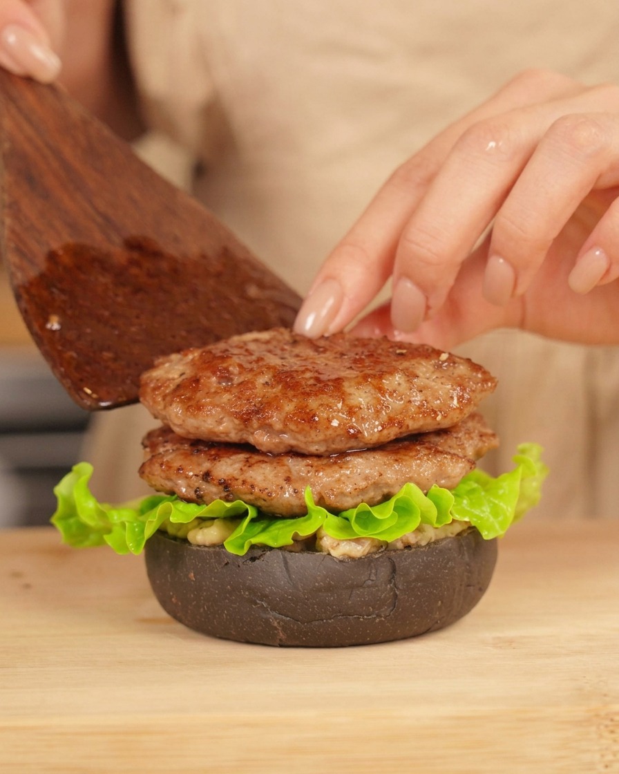 A hand using a wooden spatula to carefully place a second browned beef patty onto a partially assembled burger with a black bun and fresh lettuce.