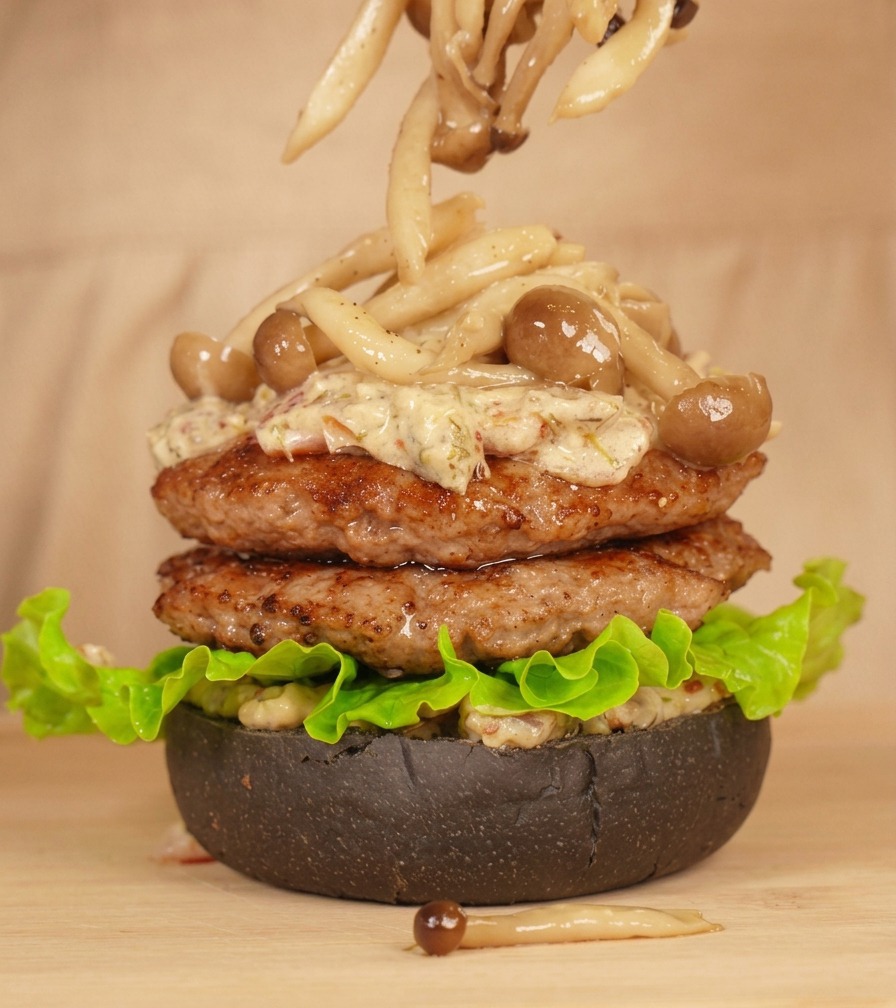 Tongs carefully placing long beech mushrooms on top of a burger stack coated in creamy sauce.