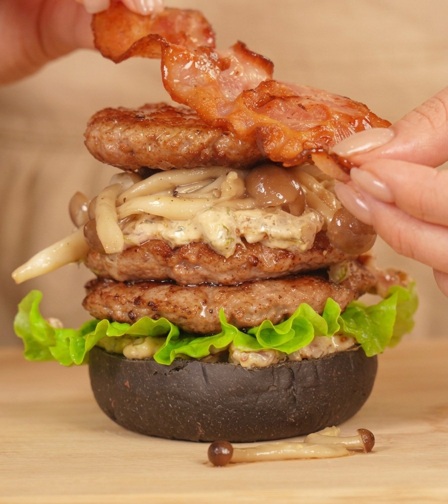 Hands placing a strip of perfectly fried bacon on top of a tall, assembled double beef burger.