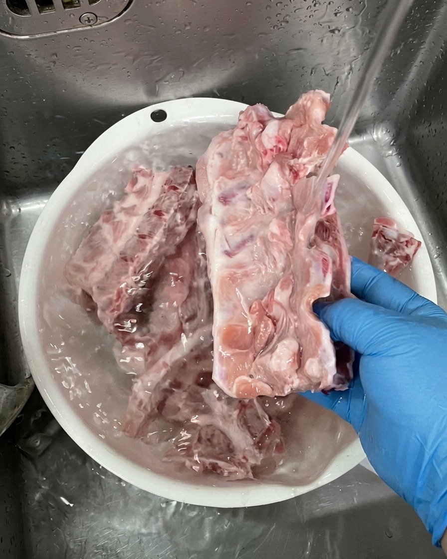 A person wearing blue gloves washing large raw pork bones under a stream of water in a white plastic basin.