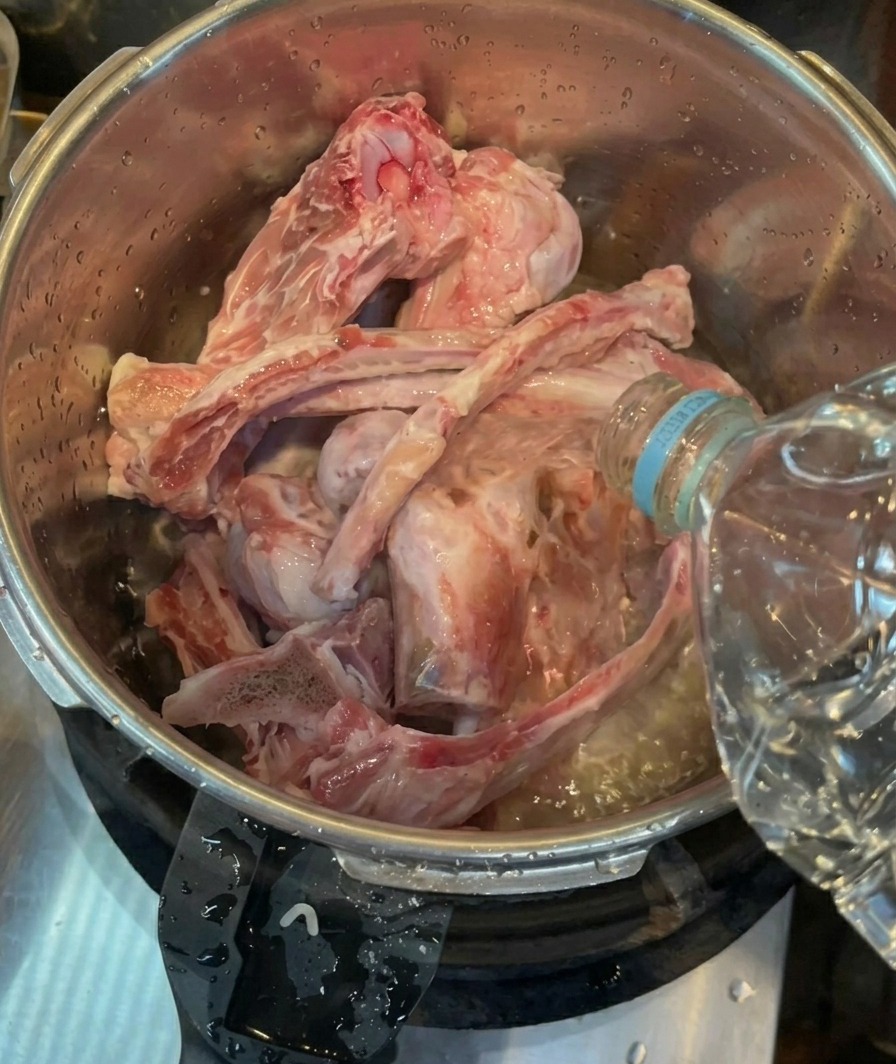 Bottled water being poured over cleaned raw pork bones inside a stainless steel pressure cooker pot.