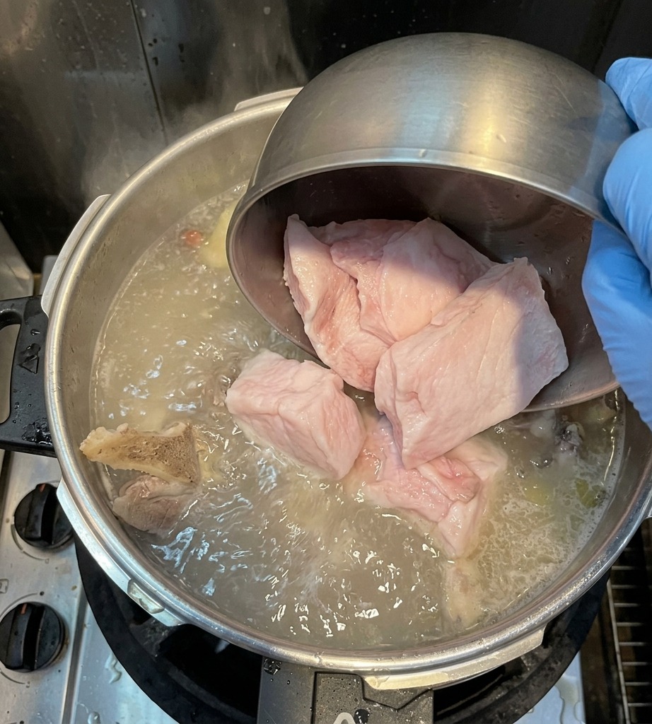 A gloved hand pouring large chunks of white pork back fat from a metal bowl into a boiling pot of broth.