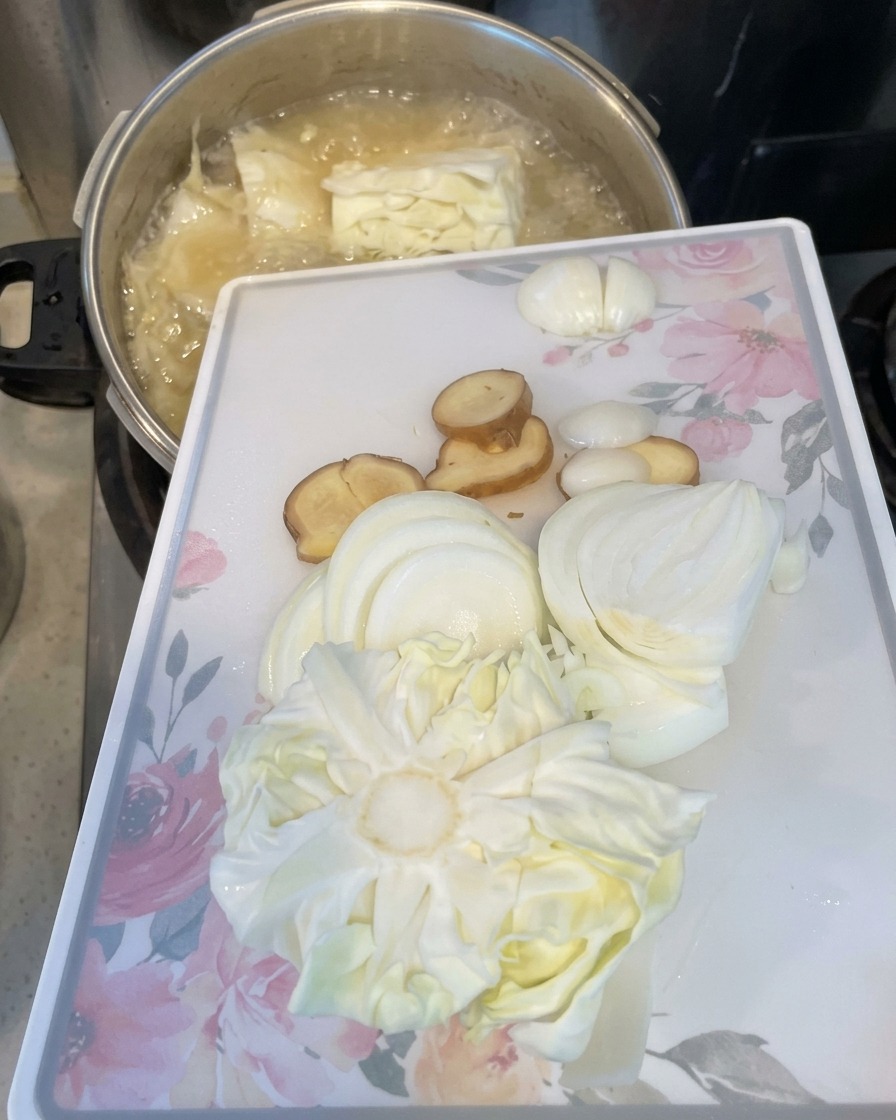 A white cutting board with sliced onions, ginger, and cabbage being tilted to slide the ingredients into a boiling pot.
