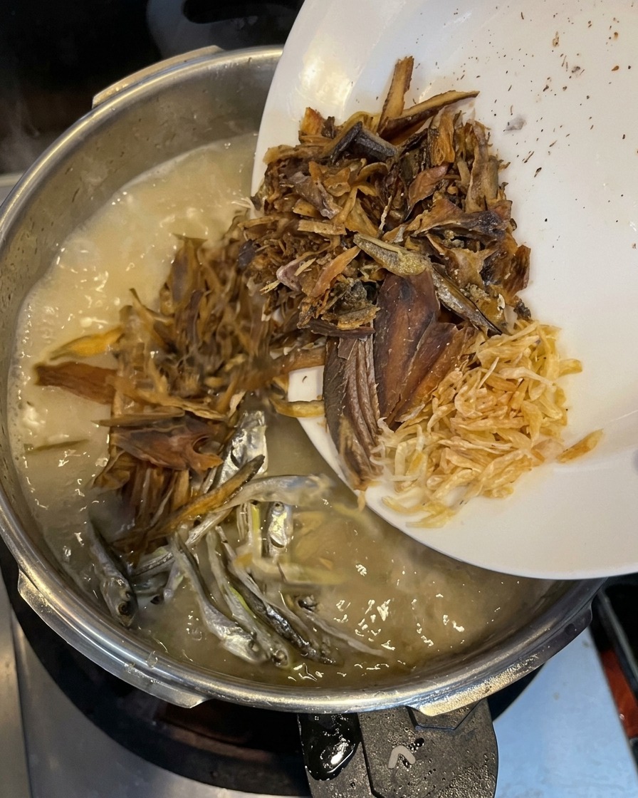 Dried fish flakes, small silver sardines, and tiny dried shrimp being poured from a white plate into a creamy boiling broth.