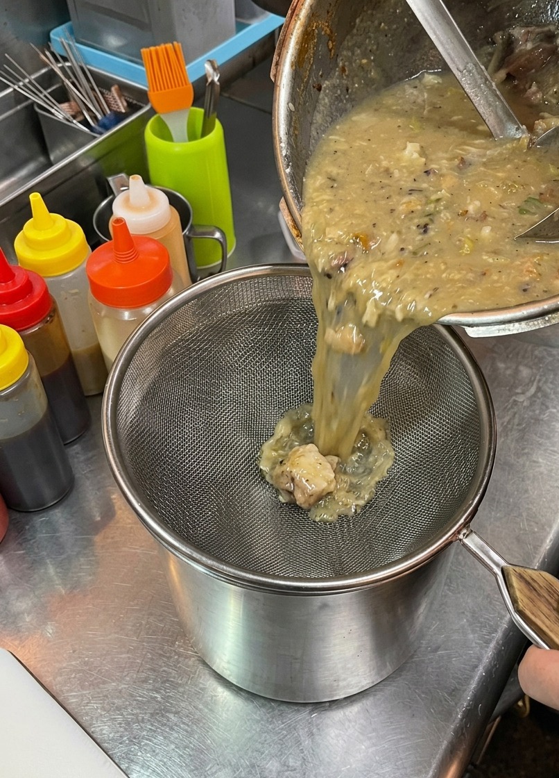 Thick, creamy ramen broth being poured from a large pot through a silver mesh strainer into a metal bin, catching pieces of pork and vegetables.