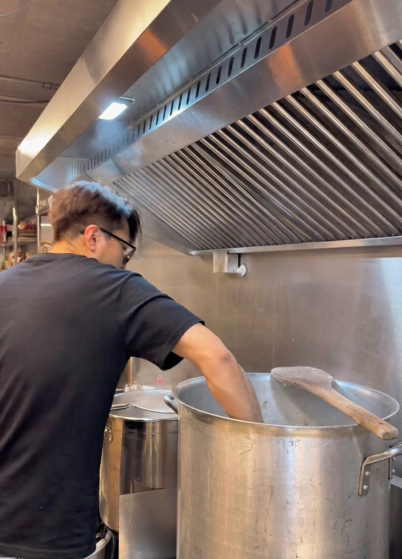 A chef in a black t-shirt leans over a large metal pot, straining a creamy white broth through a metal sieve under a industrial kitchen vent.
