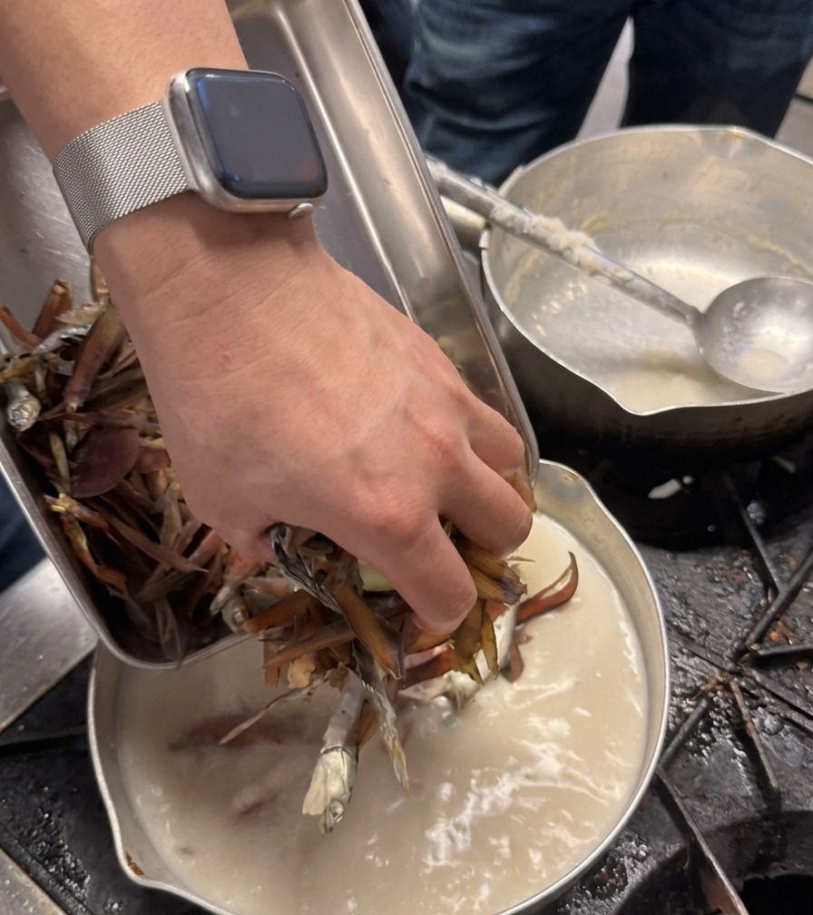 A chefs hand adding a handful of dried, dark brown ingredients into a pot of creamy white broth.