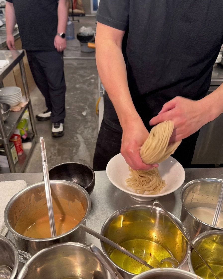 A chef using both hands to fold and press a bundle of thick, pale ramen noodles into a white ceramic bowl.