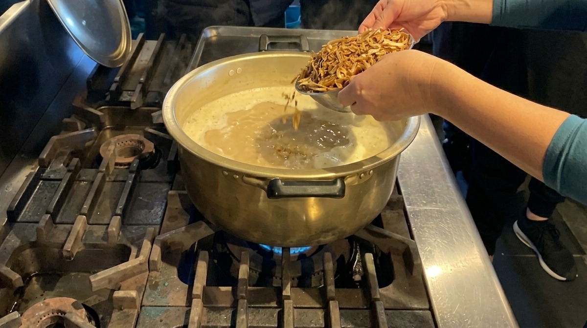 Hands adding a pile of dried anchovies into a large brass pot filled with bubbling creamy white soup.