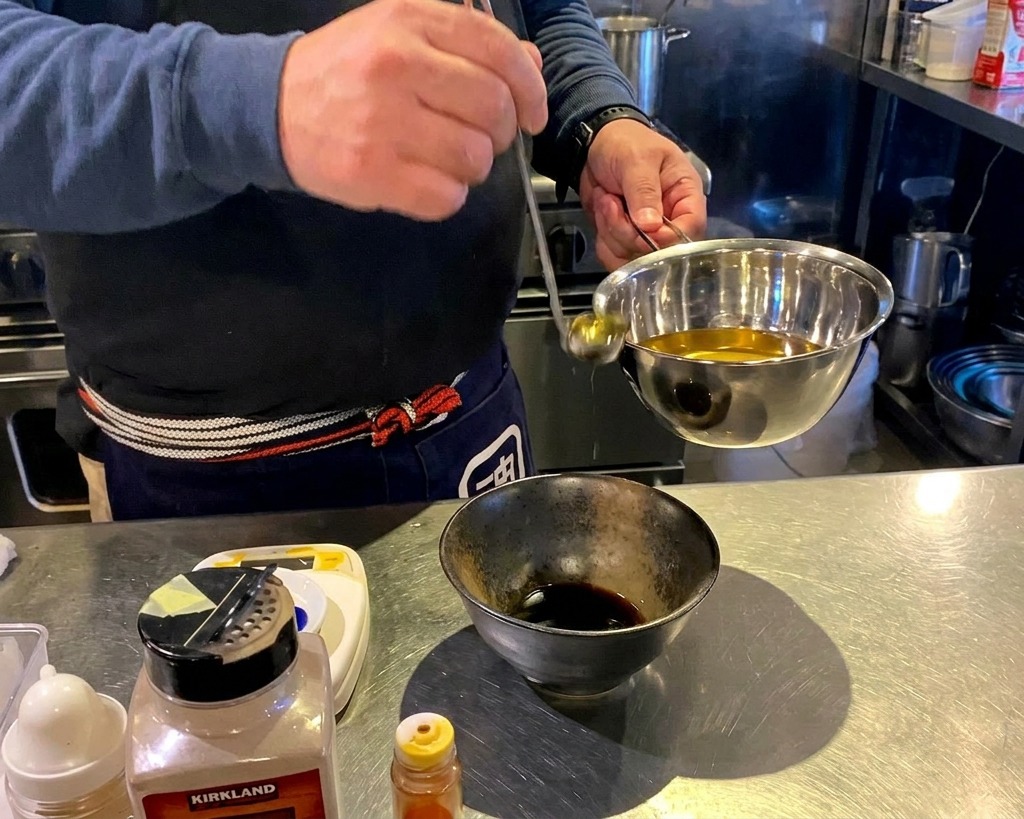 A person pouring clear golden oil from a ladle into a stainless steel bowl containing dark liquid seasoning.