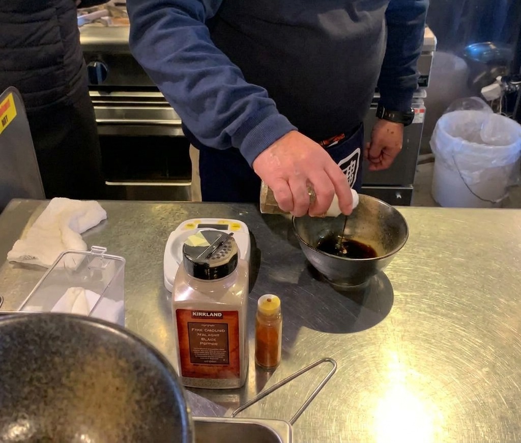 A hand pouring liquid from a bottle into a bowl that already contains a small mound of fine brown powder.