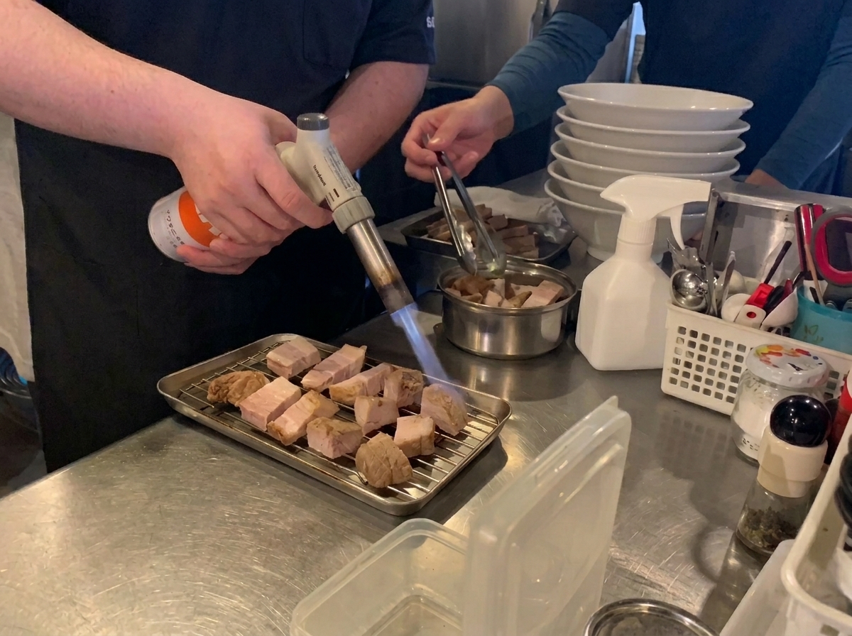 A chef using a butane torch to char and caramelize slices of braised pork belly on a metal cooling rack.