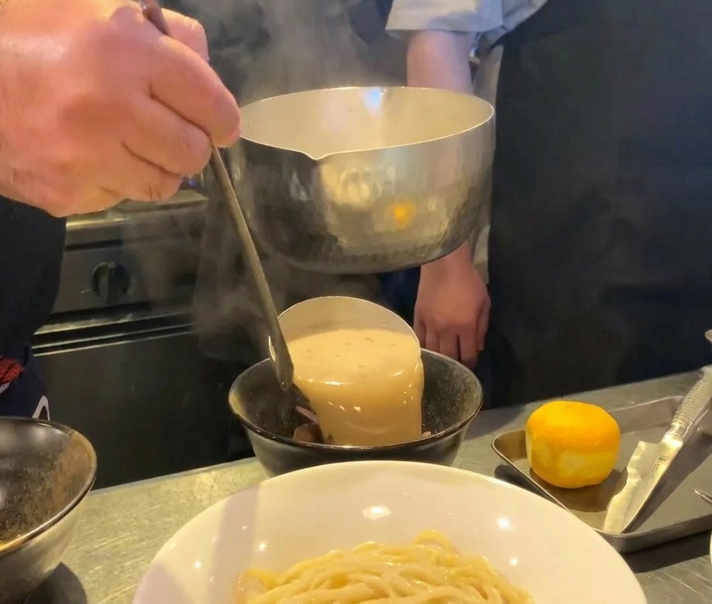 Thick, creamy seafood ramen broth being poured from a metal ladle into a black ceramic bowl.