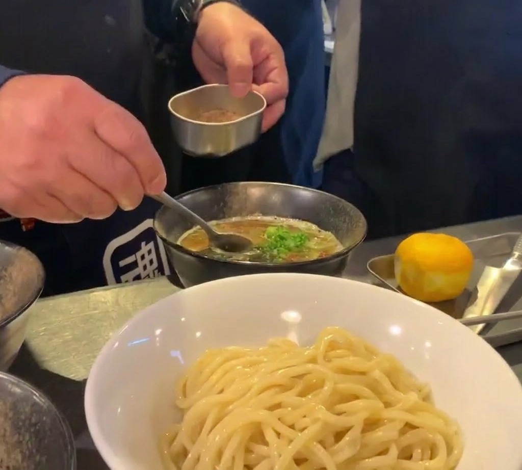 A chef using a small metal container and a spoon to sprinkle fine brown seafood powder over a bowl of dark rich dipping soup.