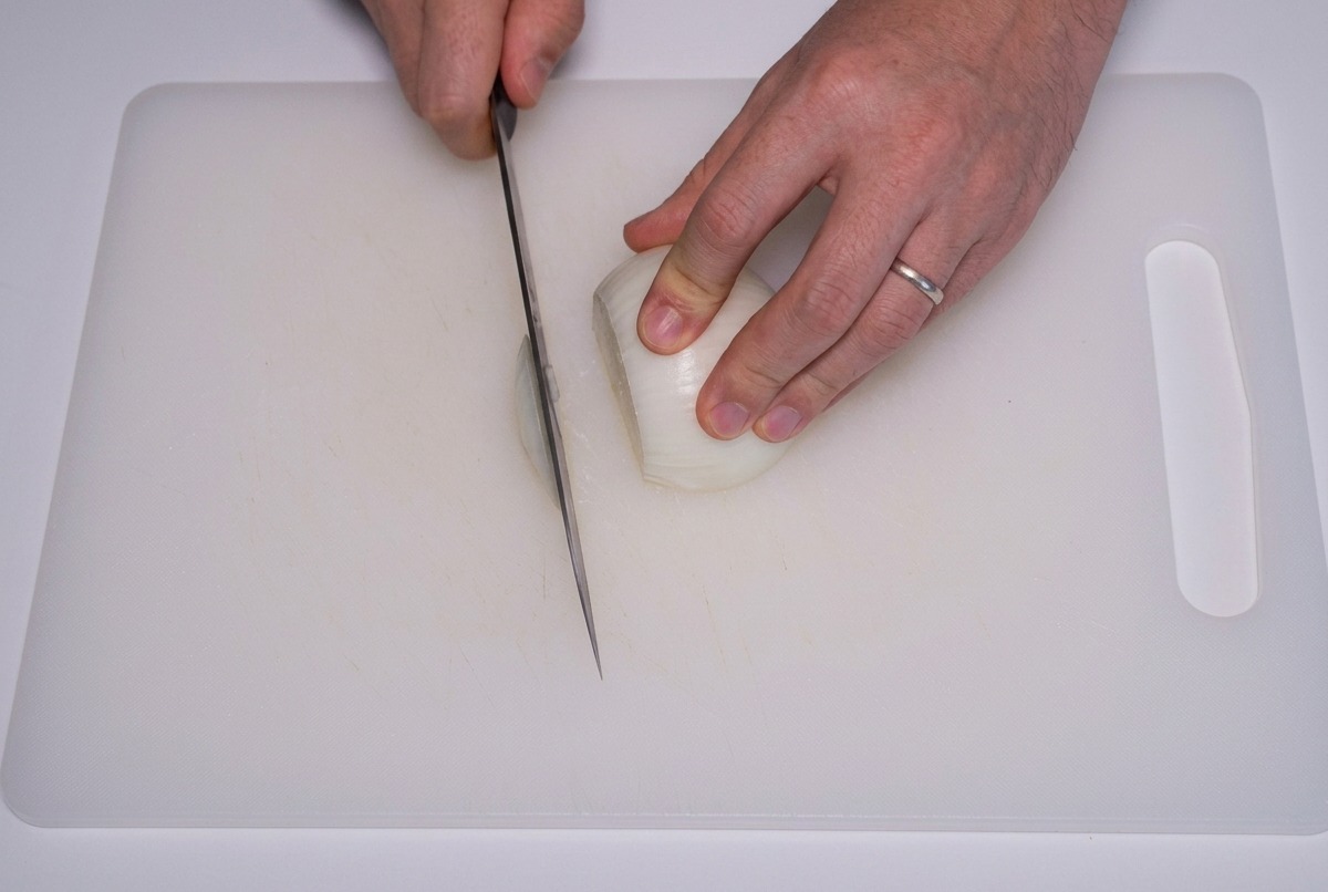 A persons hands using a sharp knife to slice a white onion into thin vertical strips on a white cutting board.