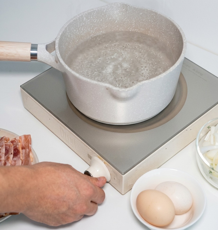 A hand turning the control knob of a white portable induction stove with a pot of water set on the heating surface.