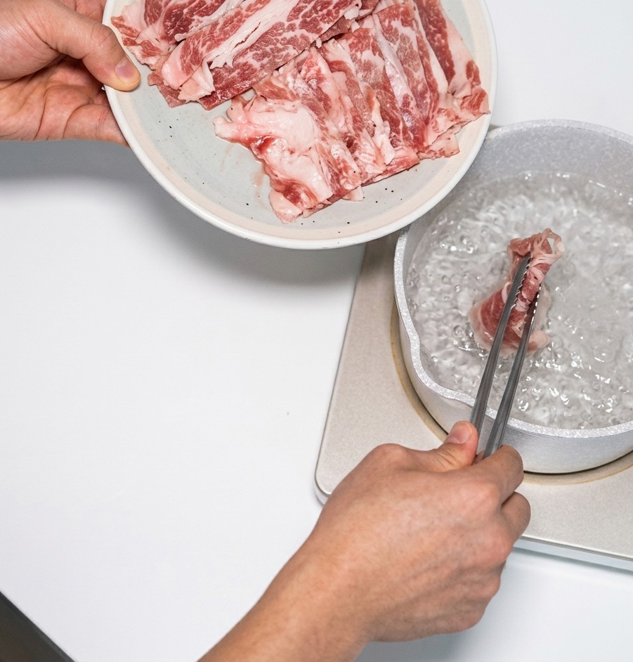 Thinly sliced marbled beef being added to a pot of boiling water using metal tongs for a quick blanch.