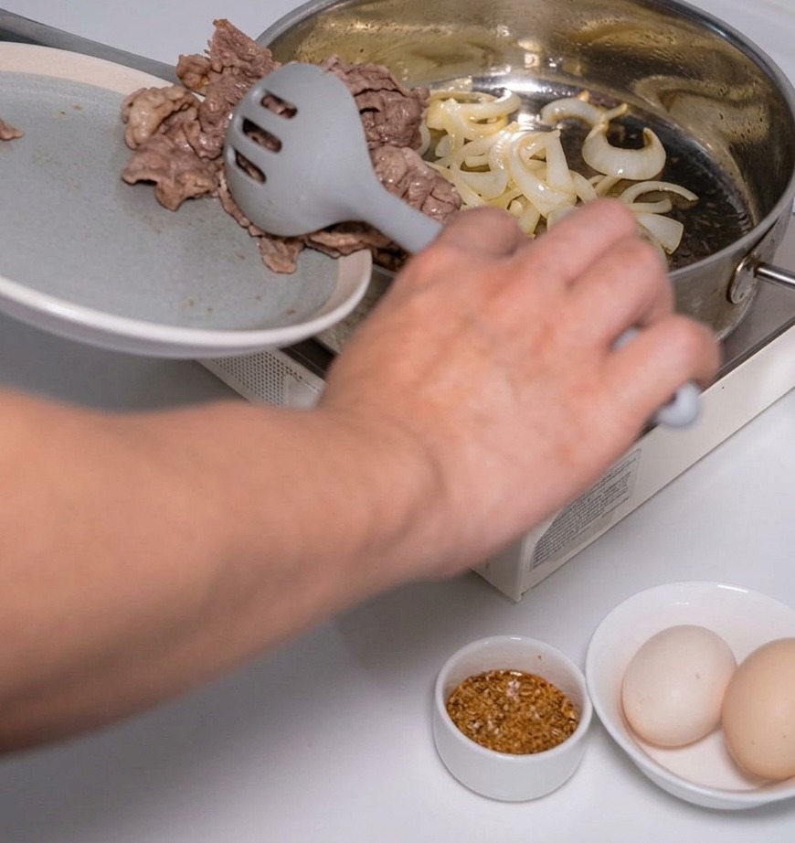 A person using a grey slotted spoon to add cooked beef slices into a pan with sautéed onions.