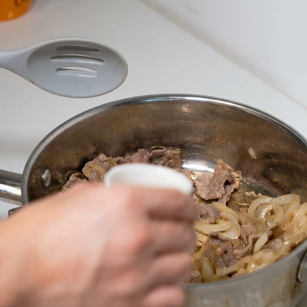 A hand pouring a small bowl of pre-mixed savory sauce over cooked beef and sliced onions in a frying pan.