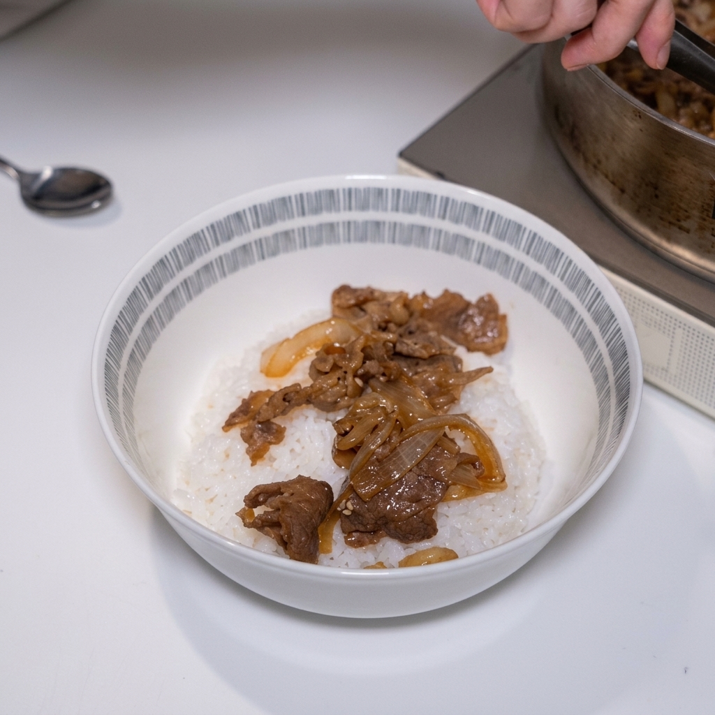 Tongs placing savory, simmered sliced beef and soft onions over a bed of steamed white rice in a bowl.