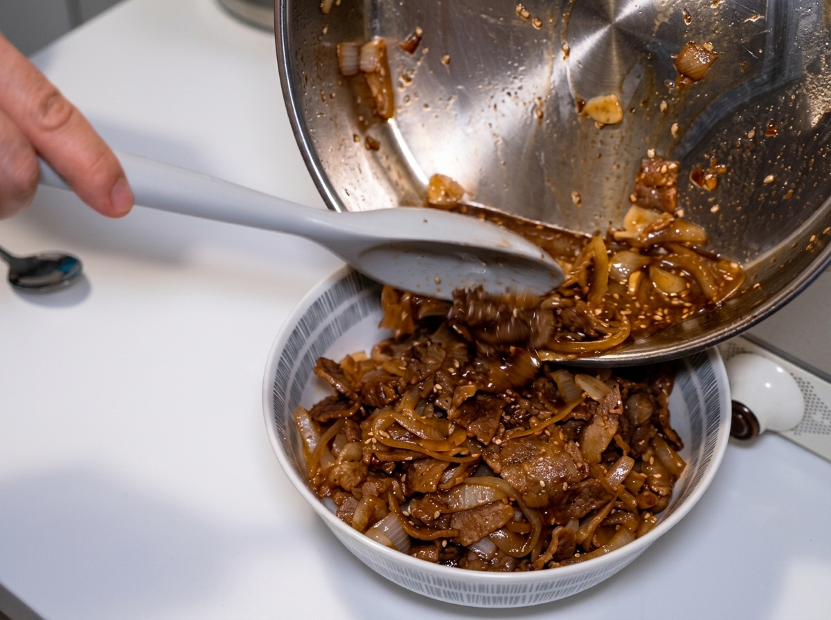 Tilting a frying pan and using a spatula to pour the remaining simmered onions and rich dark sauce over a beef rice bowl.
