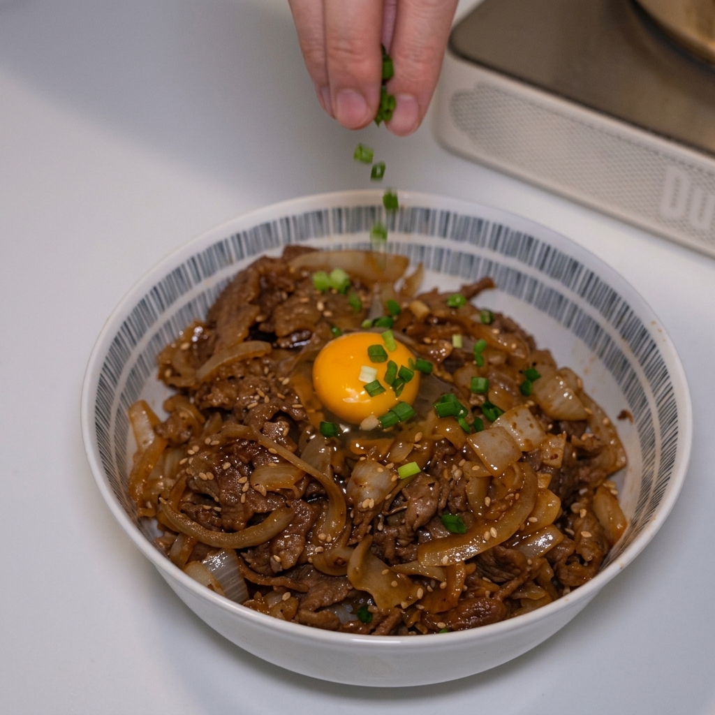 Fingers sprinkling bright green chopped scallions onto a finished Japanese beef bowl topped with a raw egg yolk.