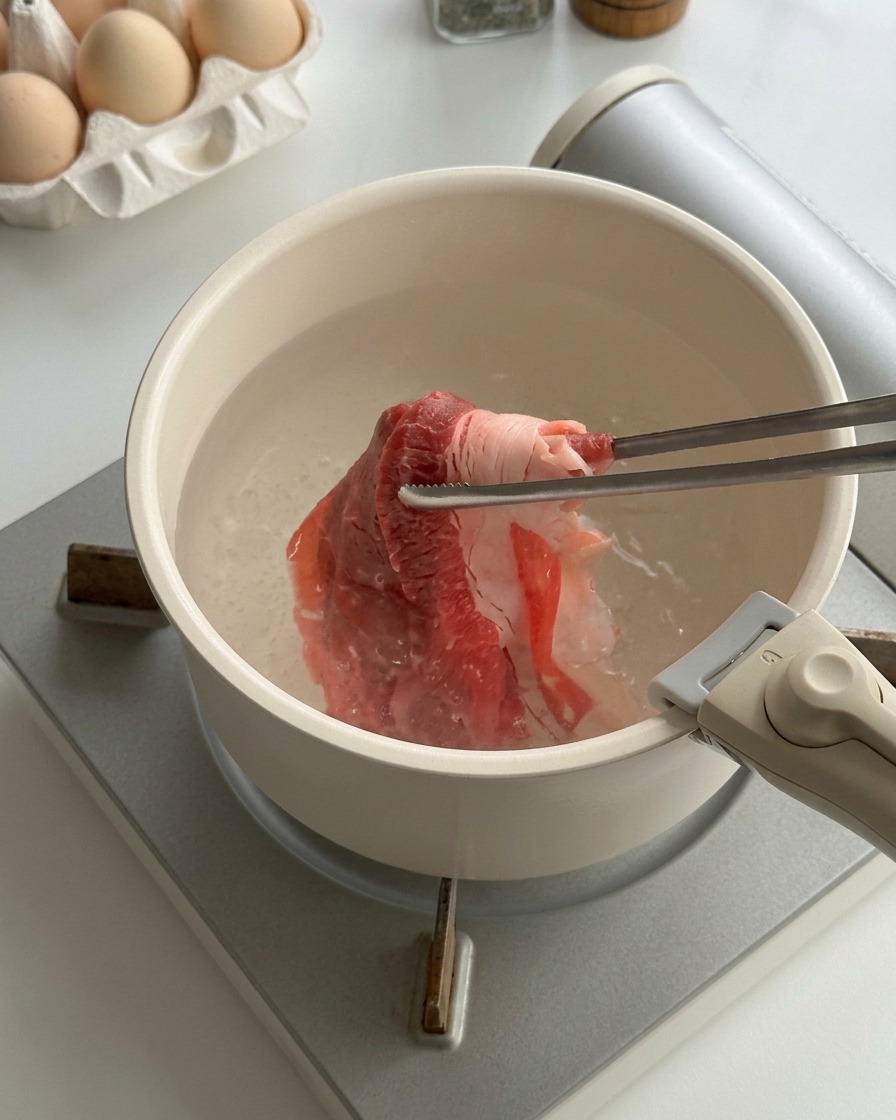 Thinly sliced raw beef being blanched in a pot of boiling water with metal chopsticks.