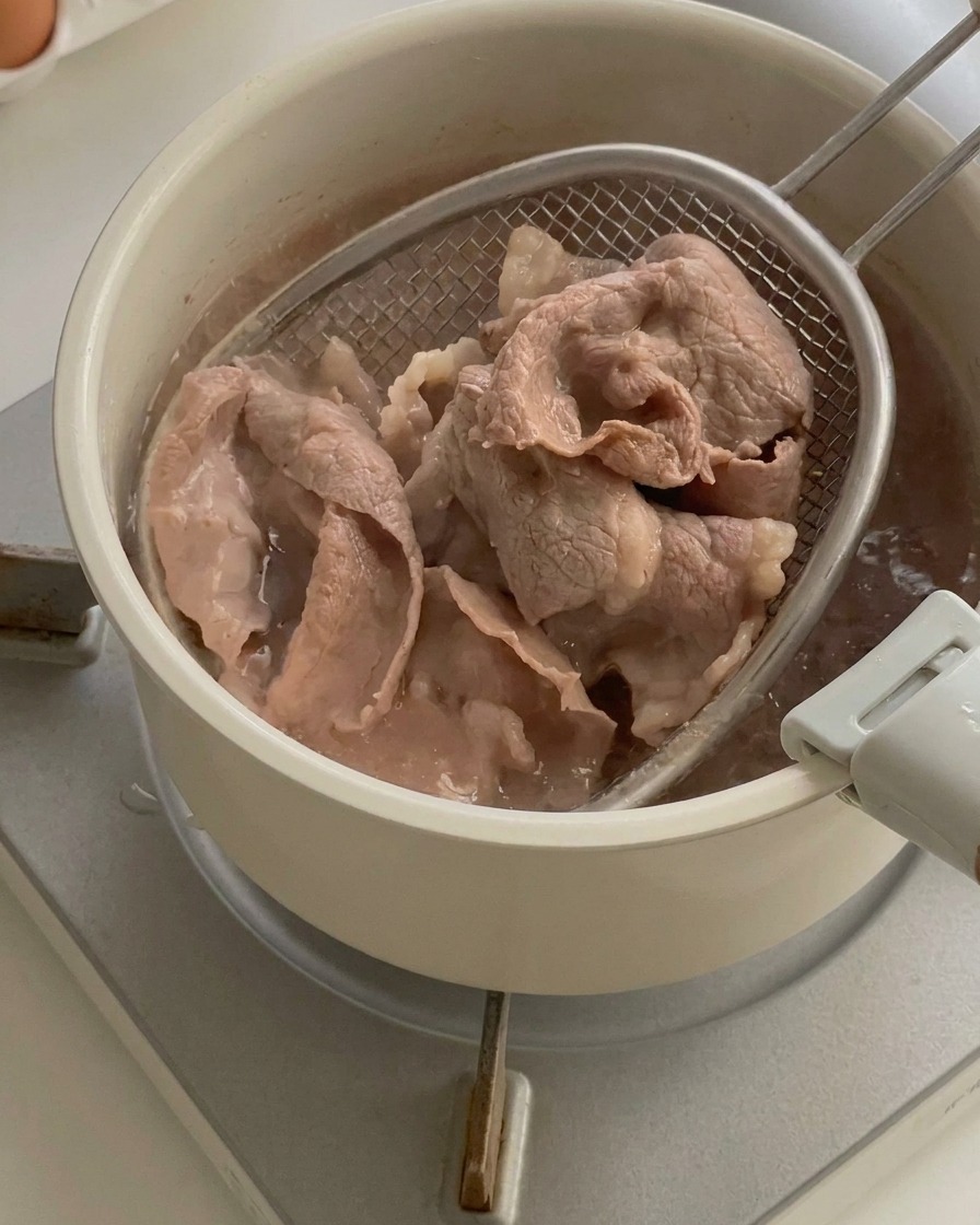 Cooked beef slices being lifted out of a pot of boiling water using a wire skimmer.