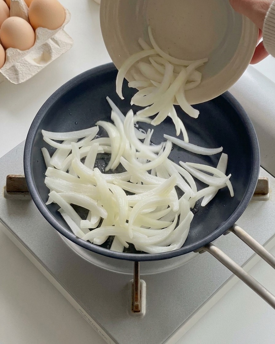 Sliced white onions being poured from a small bowl into a heated black frying pan.