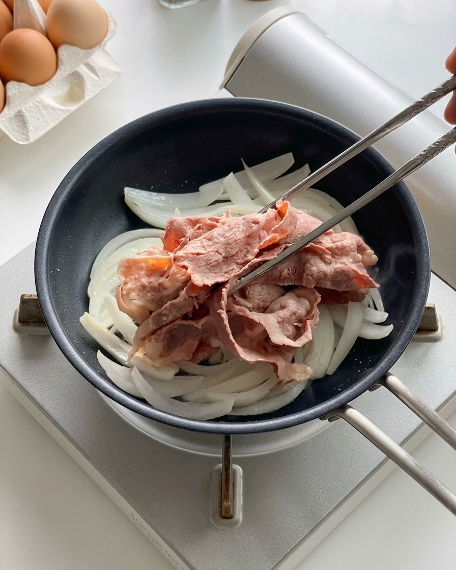 Metal chopsticks placing blanched beef slices on top of sliced onions cooking in a black frying pan.