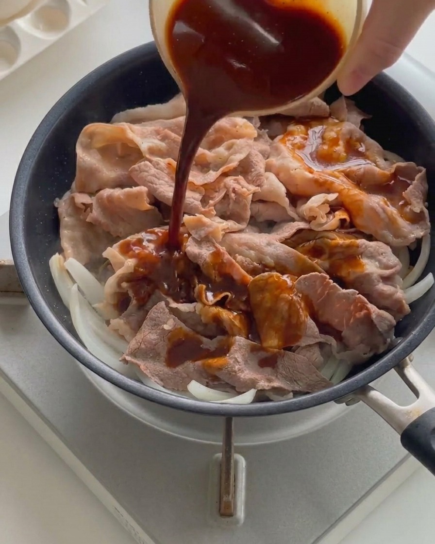Dark liquid sauce being poured from a small bowl over cooked beef slices and onions in a hot frying pan.