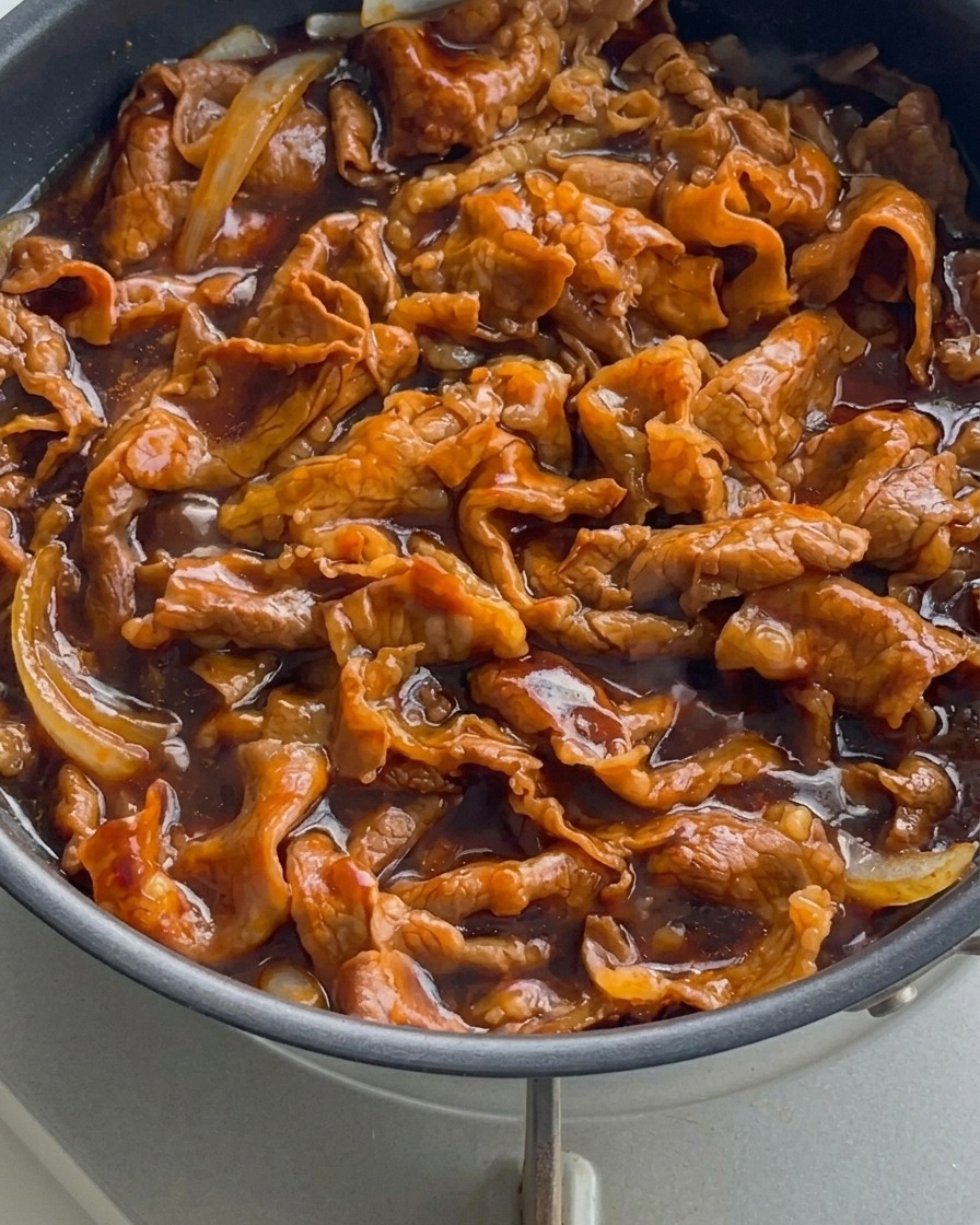A close-up view of thinly sliced beef and onions bubbling in a thick, dark, glossy sauce in a skillet.