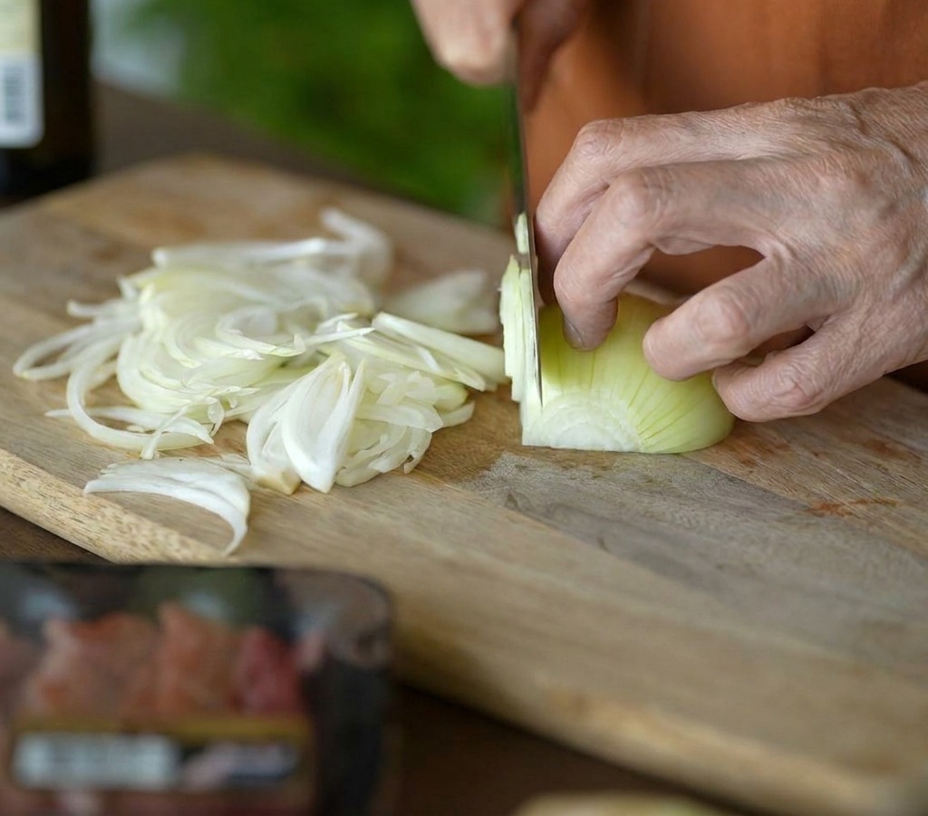A person using a sharp knife to thinly slice a yellow onion into strips on a wooden board.