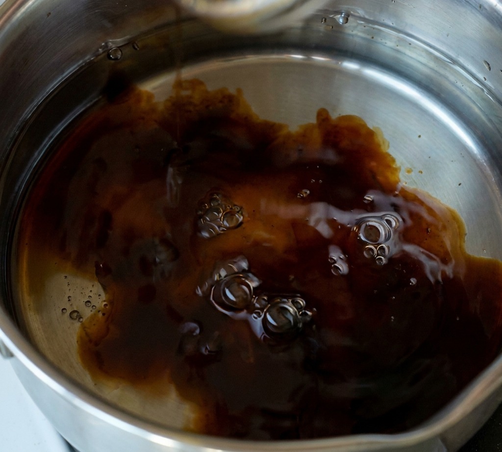 A hand adding a spoonful of white granulated sugar into a stainless steel pot filled with a dark, simmering soy-based sauce.