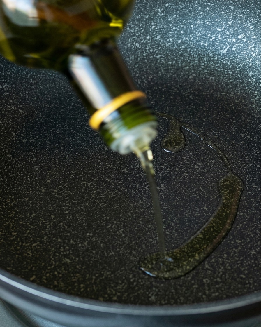 Clear avocado oil pouring from a green glass bottle onto a dark, speckled non-stick frying pan.