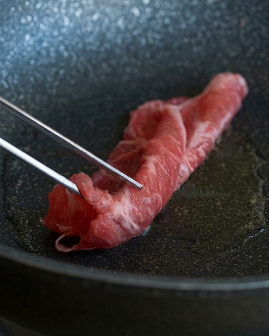 Metal tweezers placing a thin, marbled slice of raw red beef into a hot pan with shimmering oil.