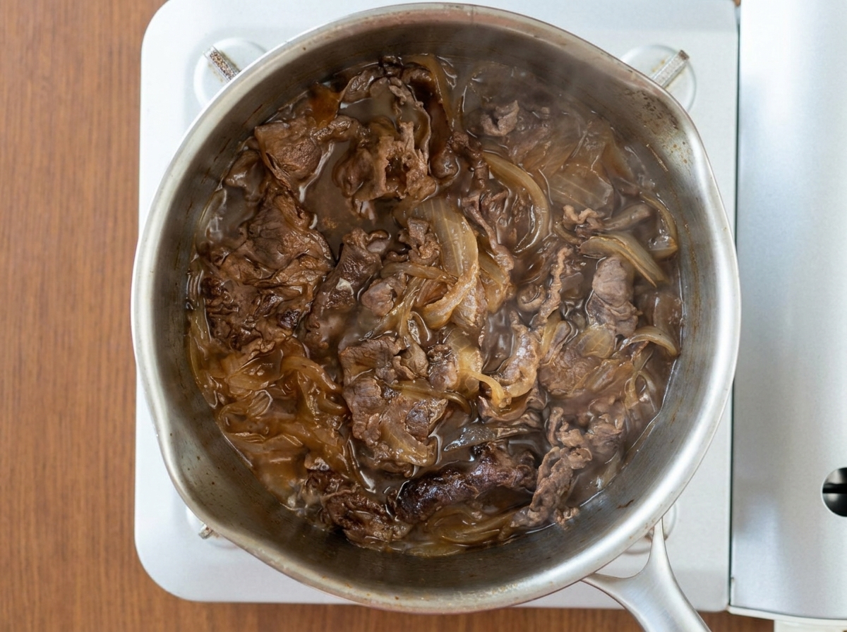 An overhead view of beef slices and onions simmering together in a dark, bubbling sauce inside a stainless steel pot.
