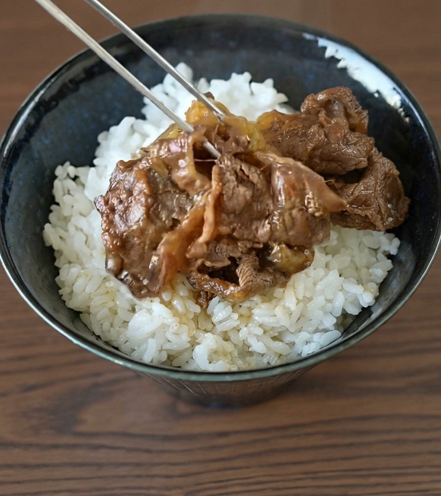 Metal chopsticks layering thinly sliced beef and onions onto a mound of fluffy white rice in a dark ceramic bowl.